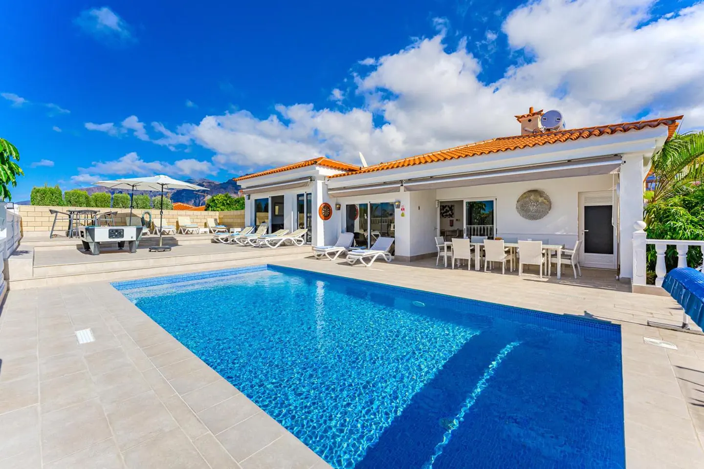 A bright blue pool sits in front of a white house with an orange tile roof, under a sunny sky with fluffy clouds. Patio furniture is visible.