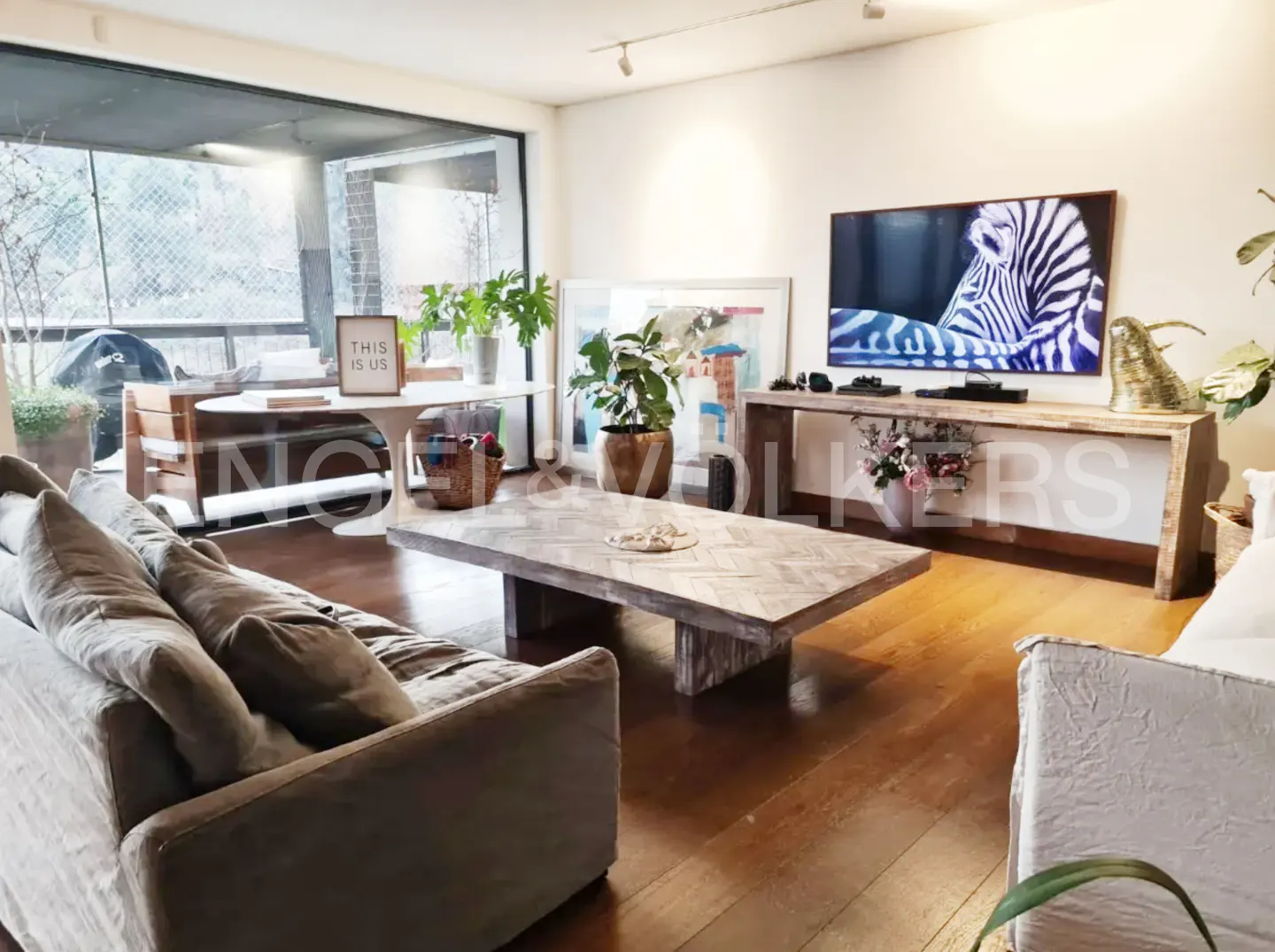 Living room with hardwood floors, a gray sofa, a herringbone coffee table, and a TV showing a zebra. A balcony is visible through a large window.