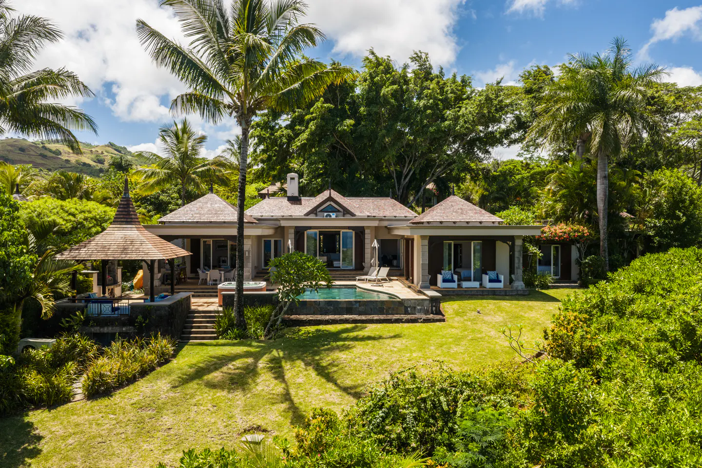 Luxury home with a pool and gazebo surrounded by lush green trees and a blue sky with white clouds.