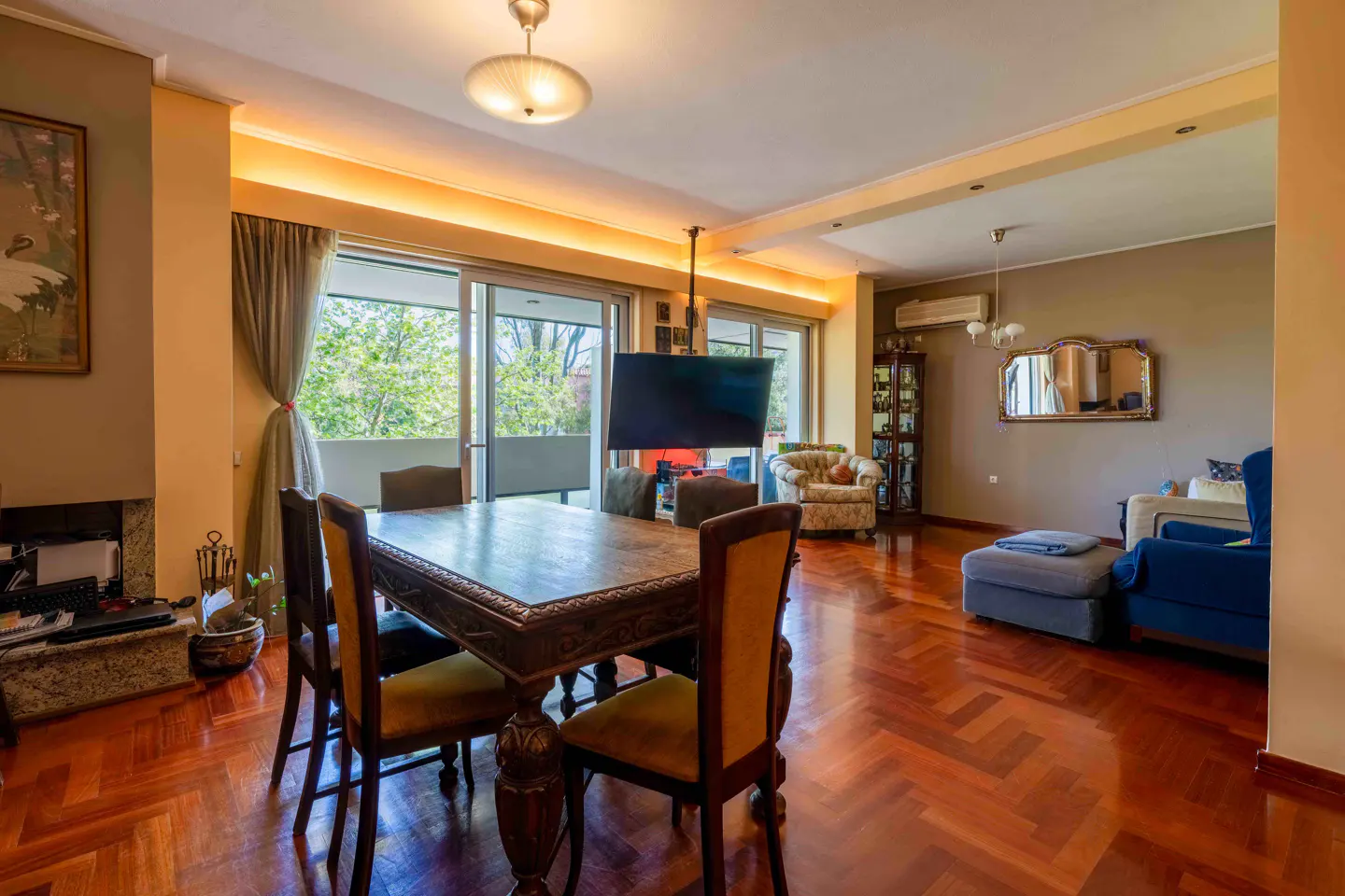 A living and dining area with a large wooden table, chairs, and herringbone wood floors. A TV hangs from the ceiling. Balcony doors let in natural light.