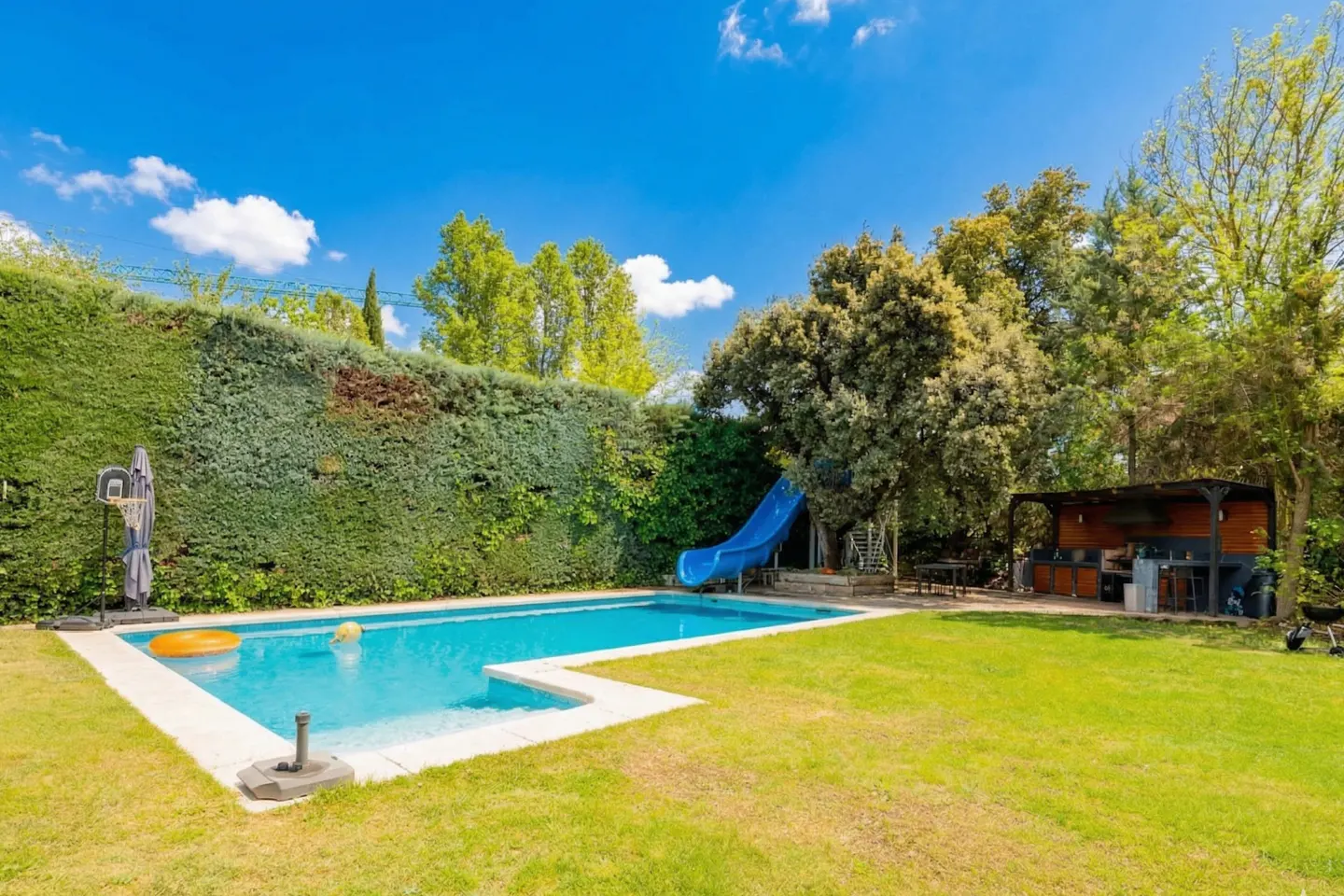 A backyard with a pool, slide, and outdoor kitchen on a sunny day. Green grass and trees surround the area.
