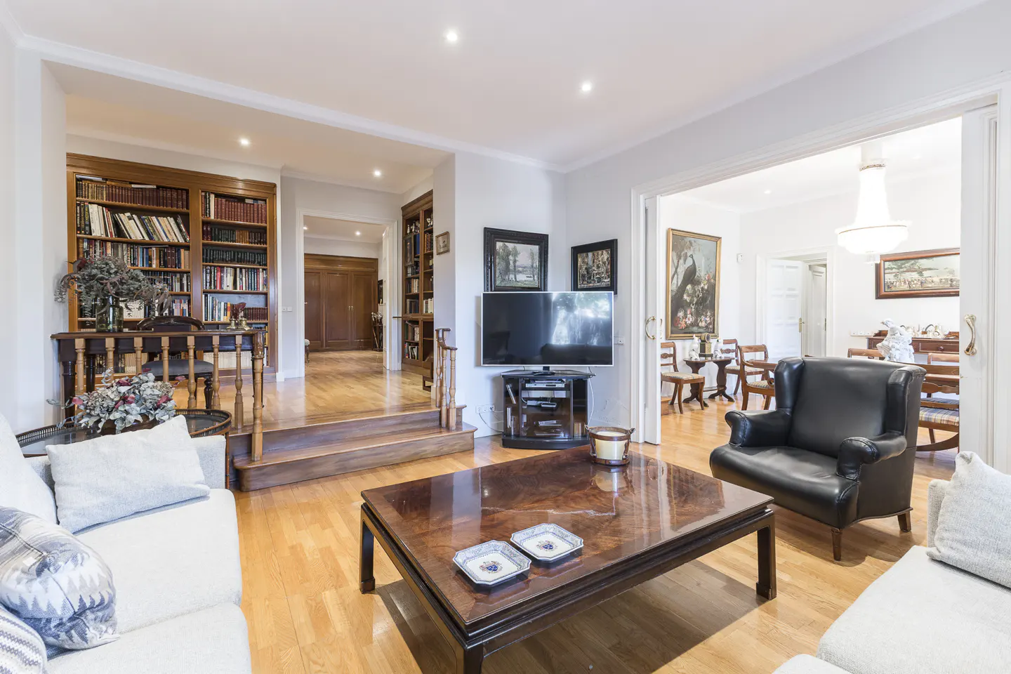 Living room with hardwood floors, white sofas, a dark wood coffee table, and a black leather armchair. Bookshelves line the walls.