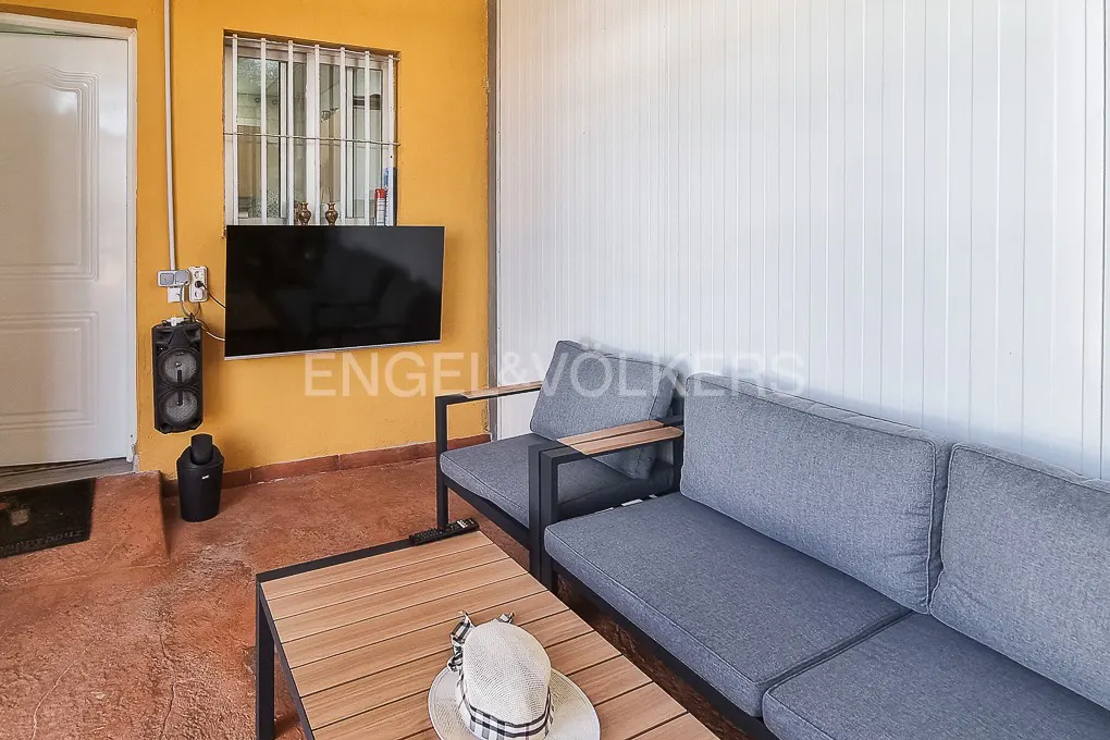 Outdoor living space with gray sofa, wood table with hat, and TV on yellow wall. White door and window are visible.