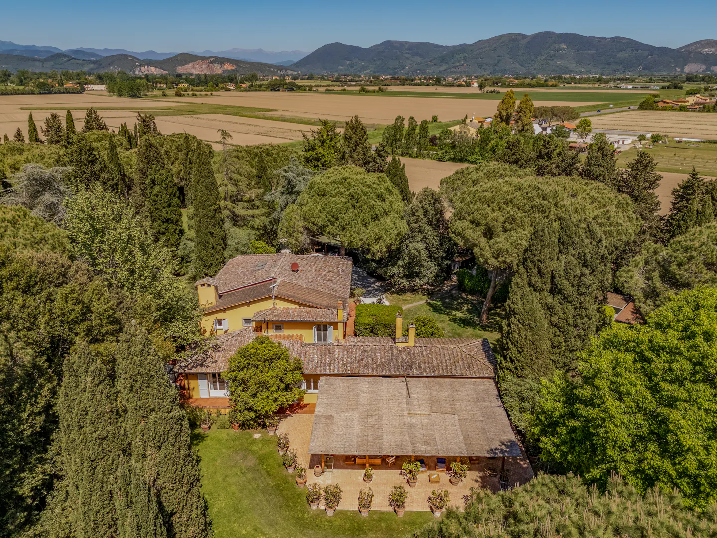 Aerial view of a yellow house with a red tile roof, surrounded by green trees and a field in the background.