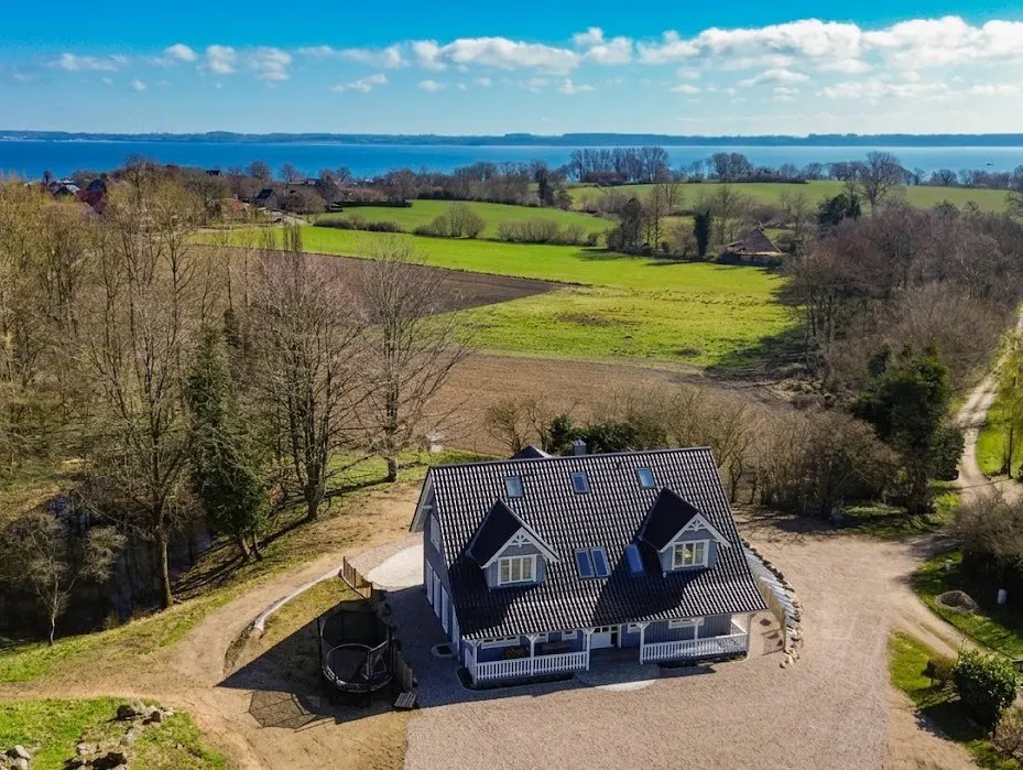 Aerial view of a gray two-story house with a black roof, white trim, and a porch, set against a backdrop of green fields and a blue bay.