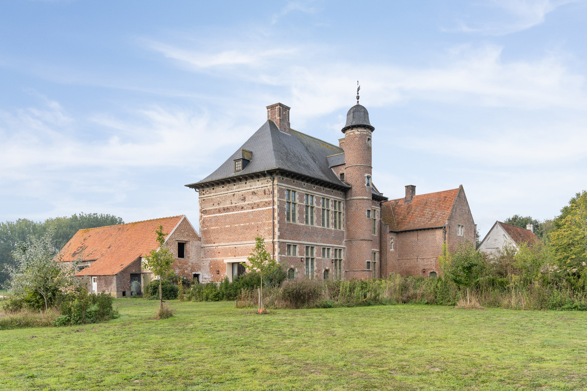 A historic brick manor house with a turret and red tile roof sits in a grassy field under a clear blue sky. [AI-generated text]