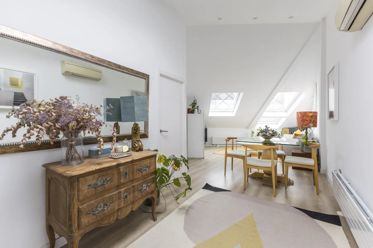 A bright attic room with a wooden dresser, a large mirror, and a dining area with a glass table and wooden chairs.