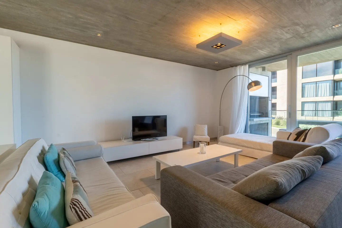 Bright living room with white walls, concrete ceiling, and large windows. Features a gray sectional sofa, white sofa, TV, and modern lighting.
