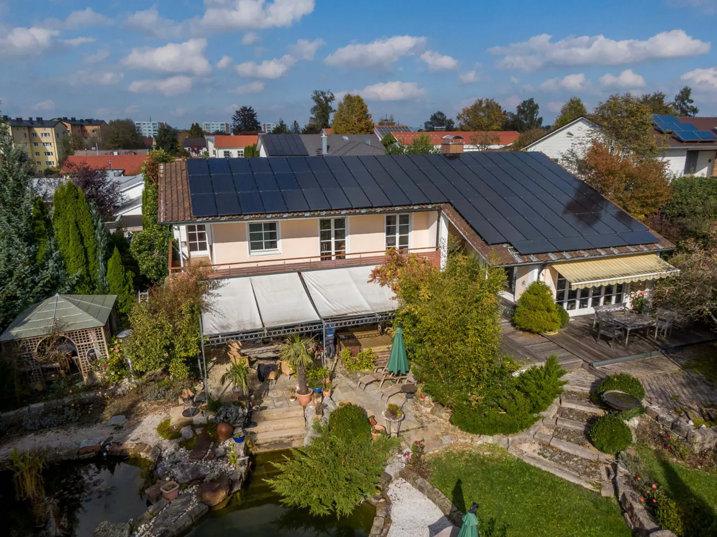 Aerial view of a two-story house with solar panels, a patio, and a pond in a lush garden.