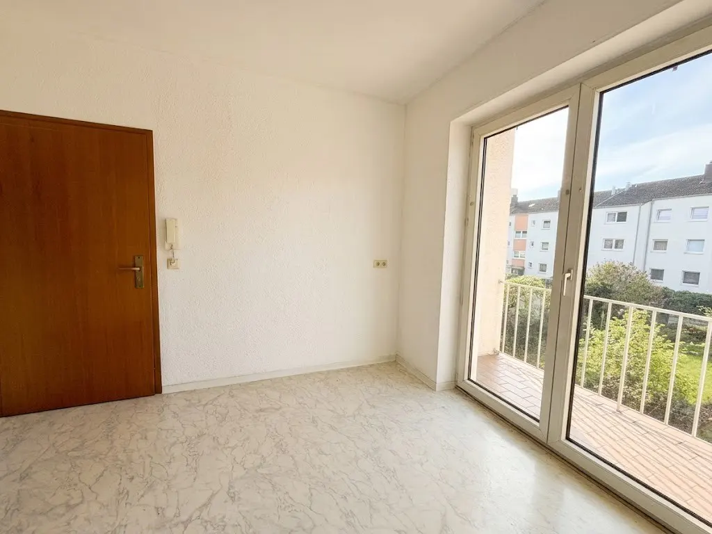 Bright, empty room with marble floor, white walls, and a wood door. Balcony with white railing visible through sliding glass doors.