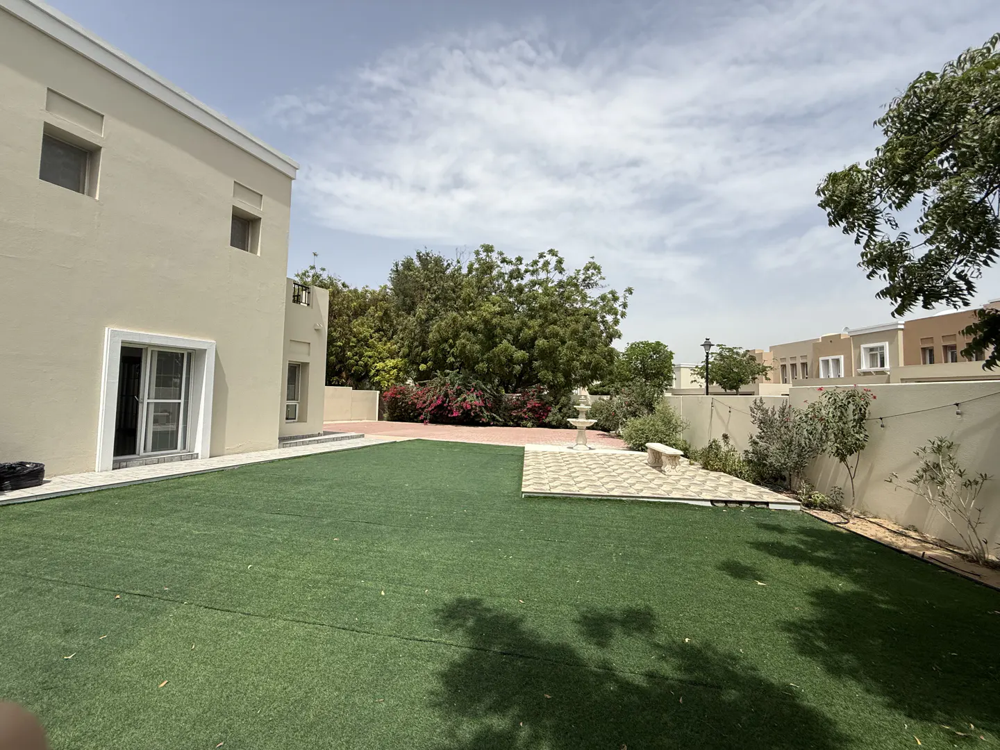 A beige house with a green lawn, a tiled patio with a fountain and a bench, and trees under a blue sky.