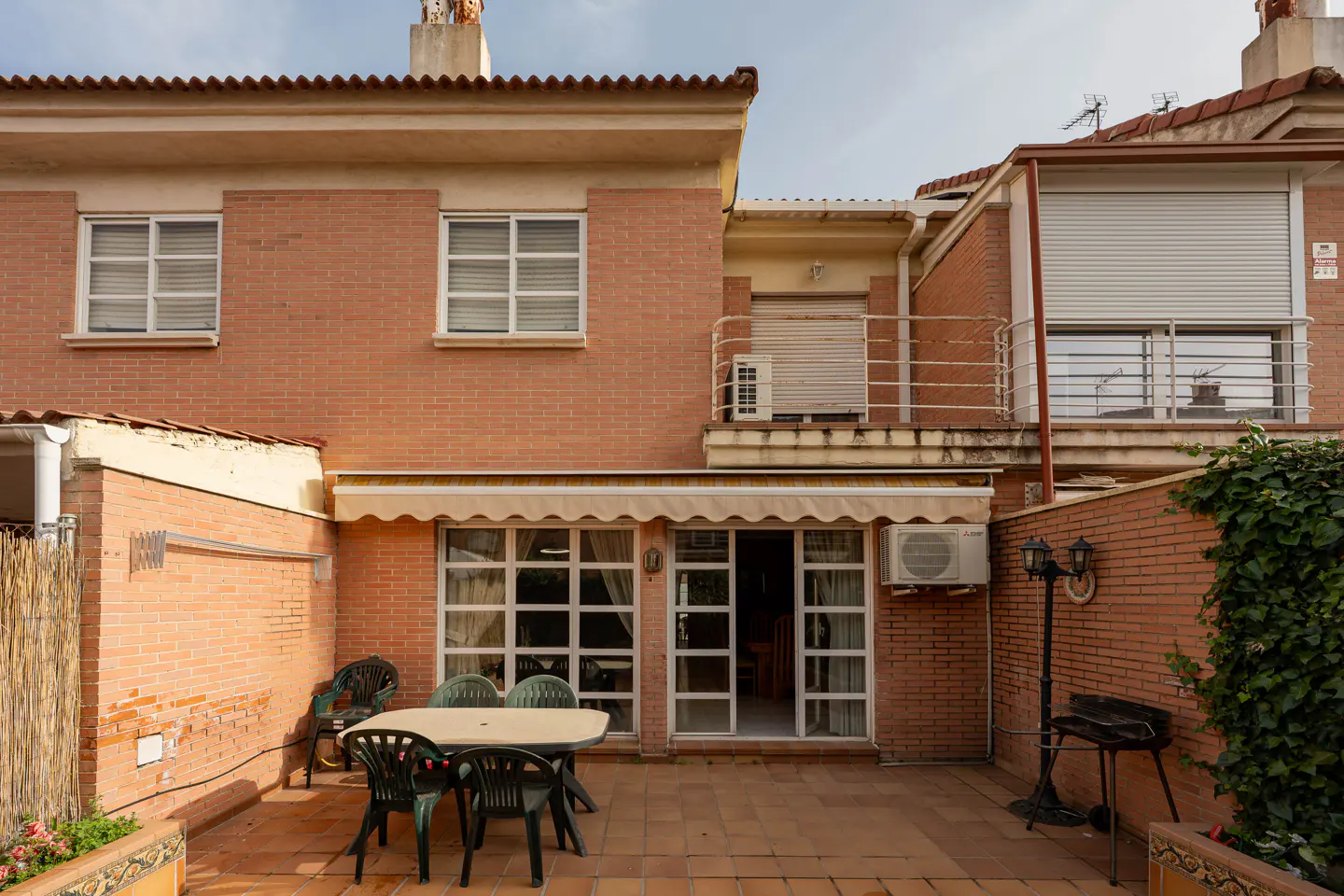 Brick townhouse exterior with a patio. Table and chairs sit on the tiled patio. A grill and plants are also visible.