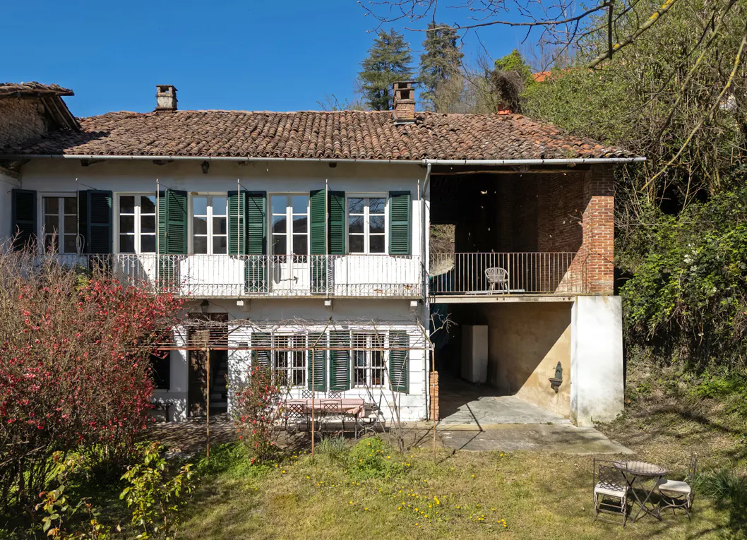 Two-story white house with green shutters, a red tile roof, and a small balcony. A lawn and trees surround the house.