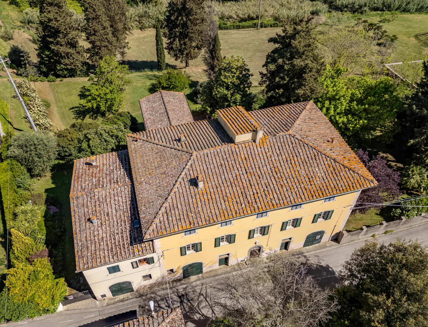 Aerial view of a yellow, two-story house with a terracotta tile roof, surrounded by green trees and grass.