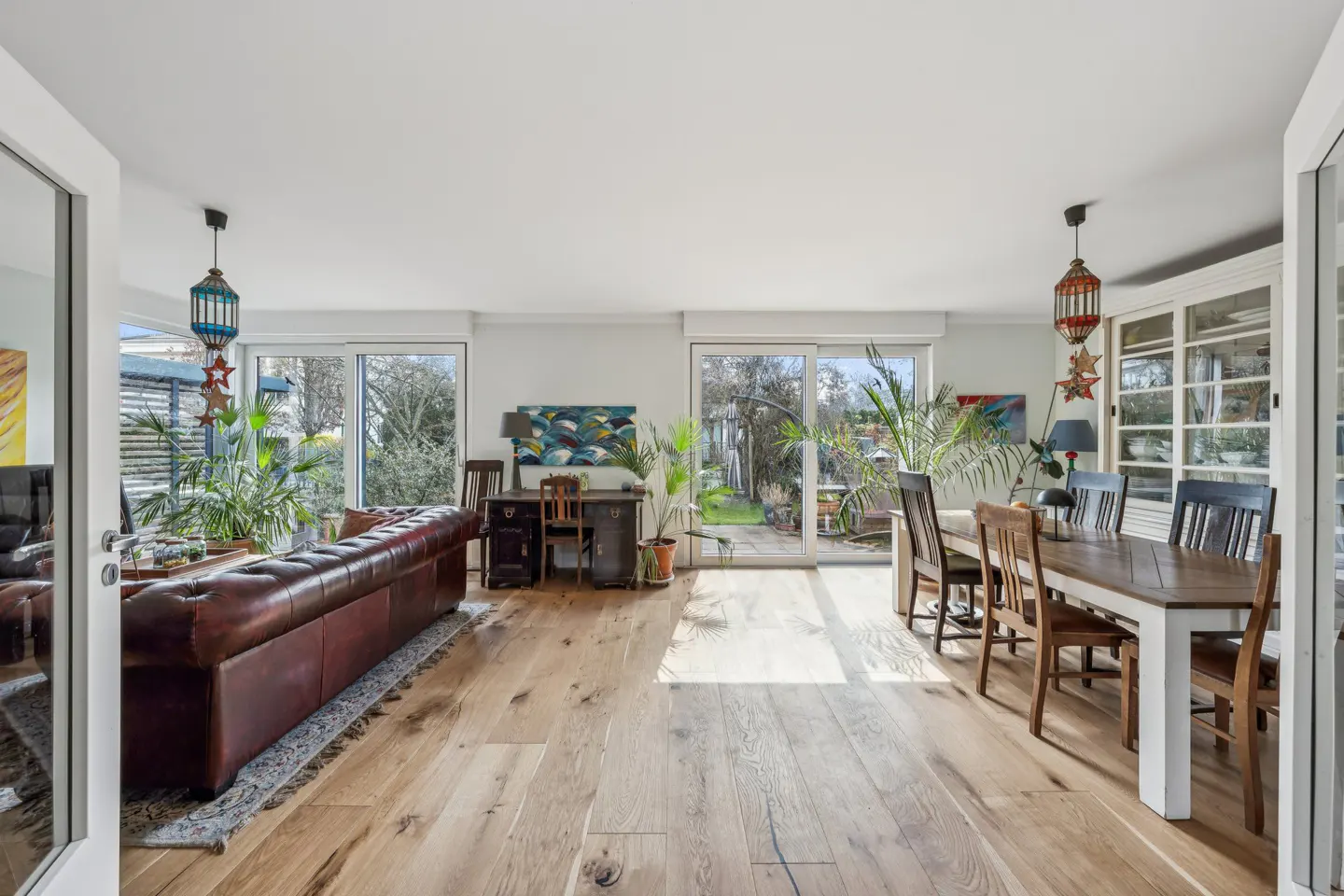 Bright living room with wood floors, a brown leather sofa, a desk, and a dining table with chairs. Sliding glass doors lead to a garden.