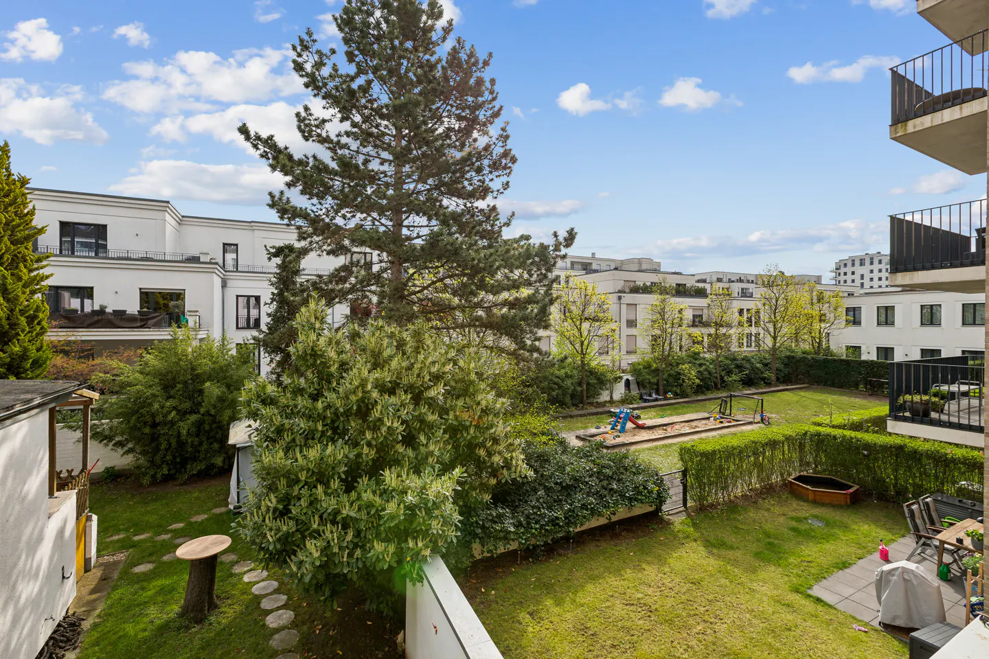 View of a green courtyard with a playground, surrounded by white buildings and trees under a blue, cloudy sky.
