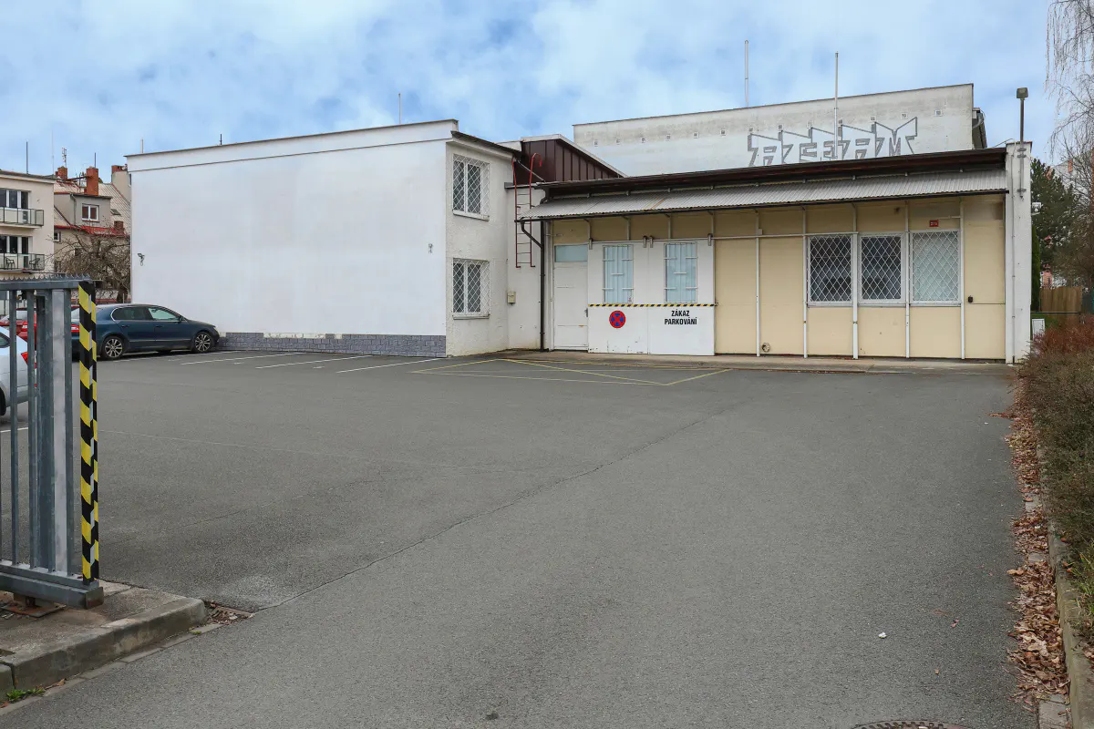 Exterior view of a white and yellow building with a parking lot in front. A blue car is parked near a metal gate.