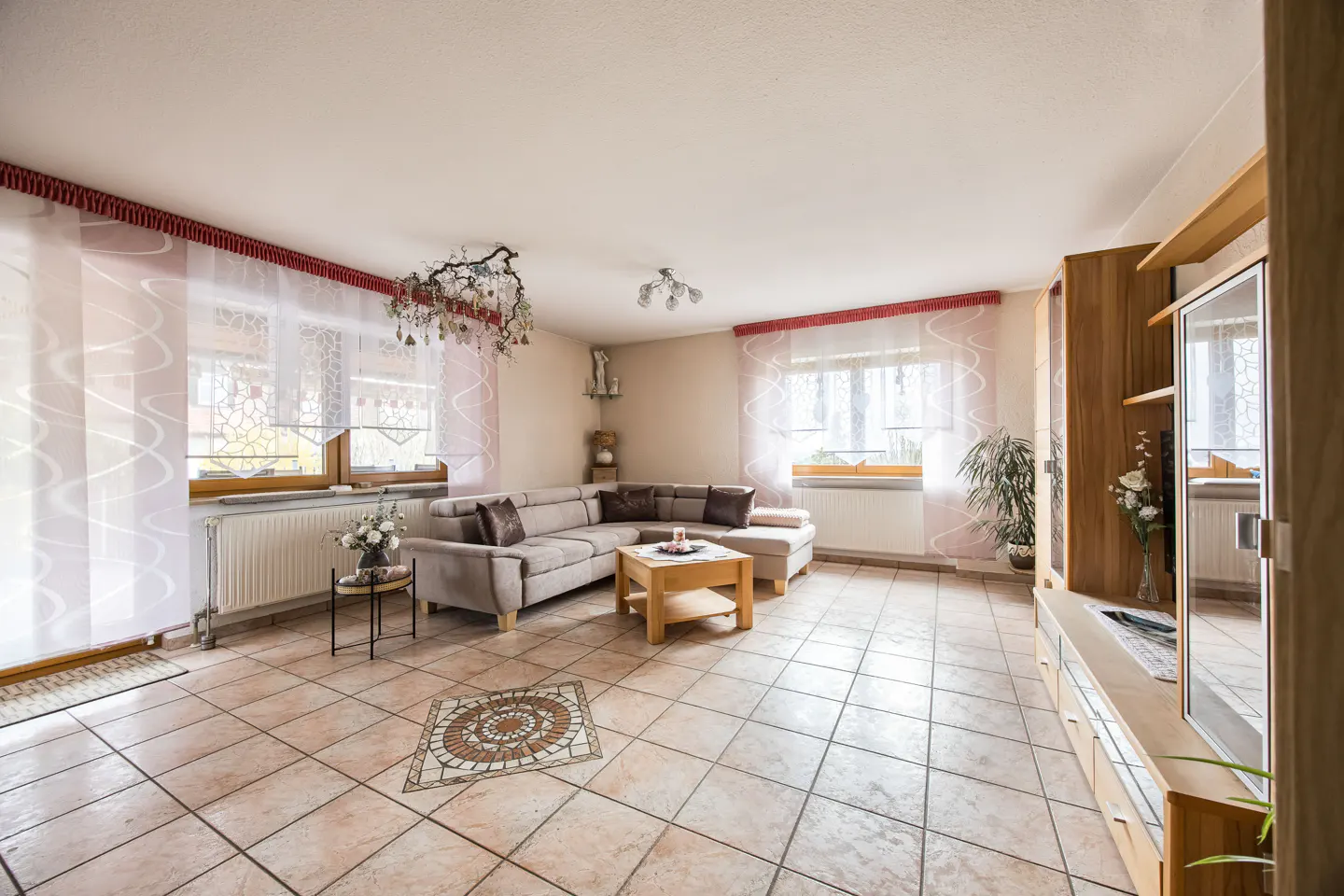 A bright living room with beige tile flooring, a sectional sofa, and sheer white curtains with red trim. A wooden entertainment center stands to the right.