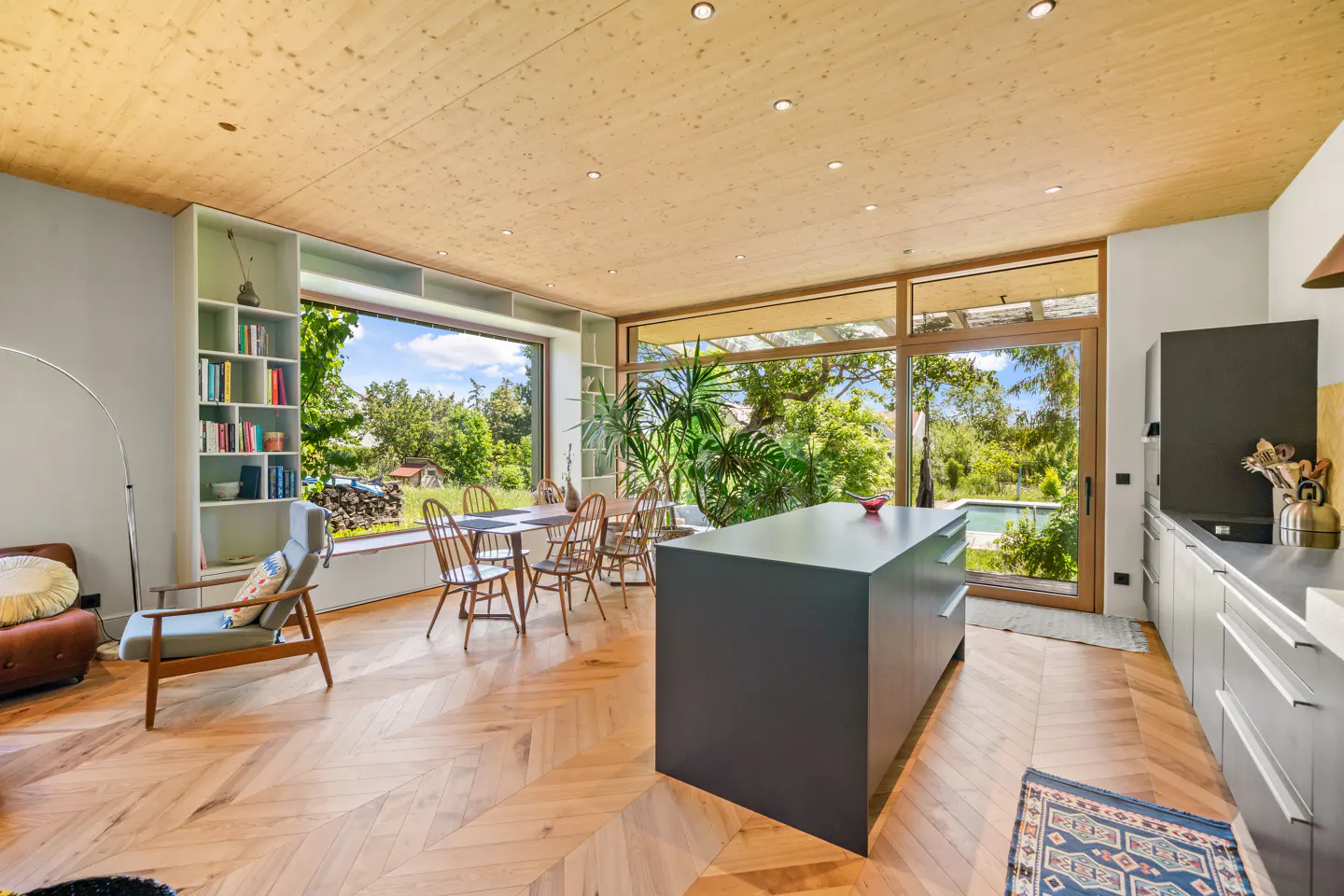 Bright, modern kitchen with wood floors and ceiling. Large windows overlook a green yard with a pool. Island, dining table, and bookshelf visible.