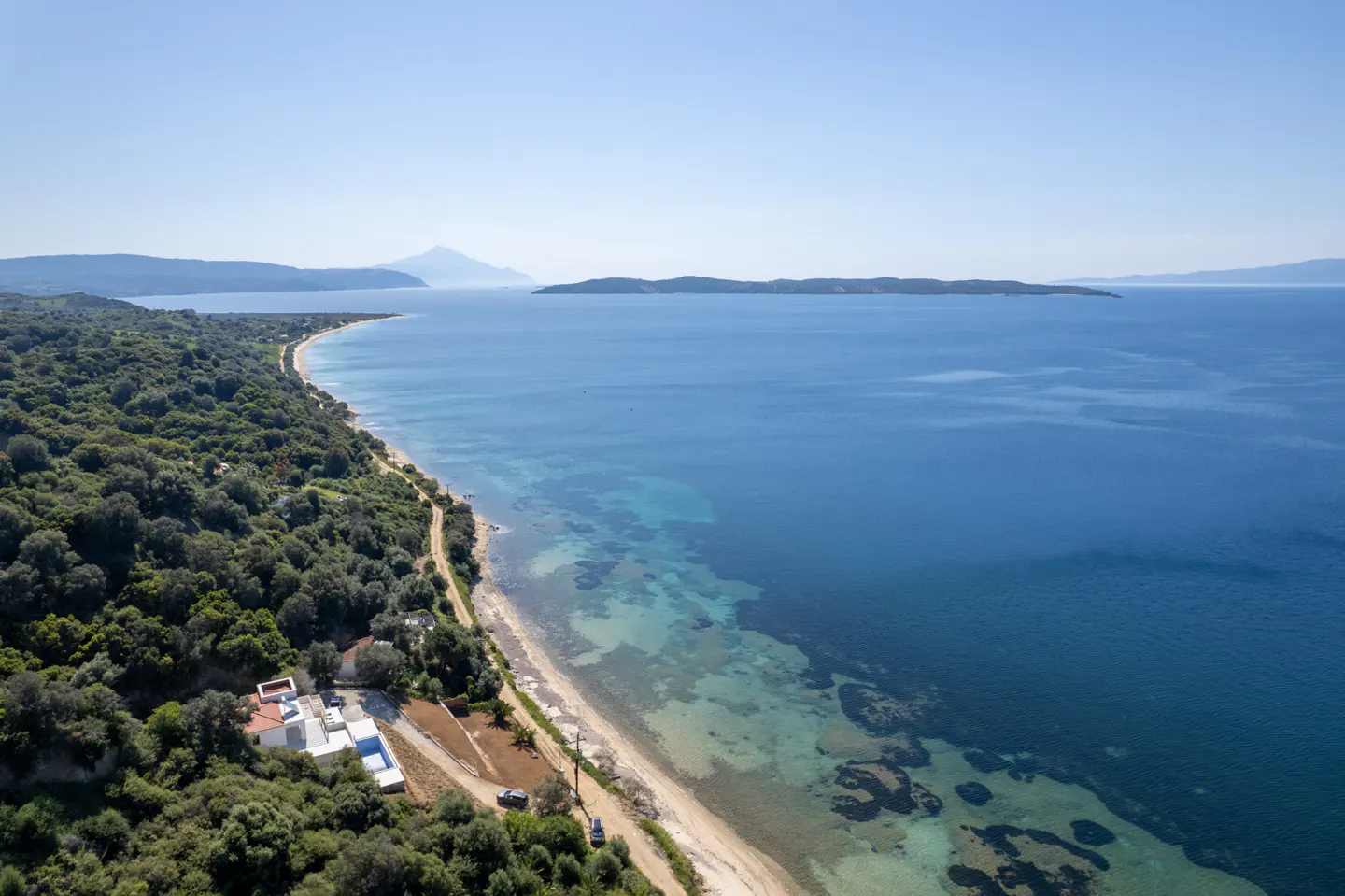 Aerial view of a white house nestled in green trees along a sandy beach and blue ocean.