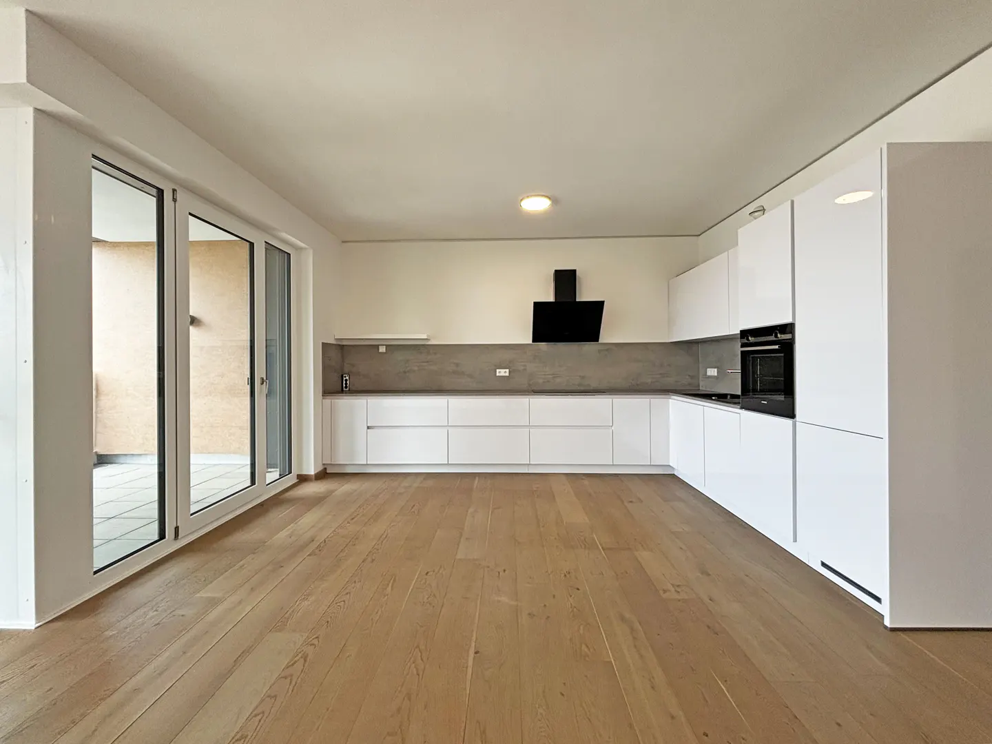 Bright, modern kitchen with light wood floors, white cabinets, and a black range hood. Sliding glass doors lead to a balcony.