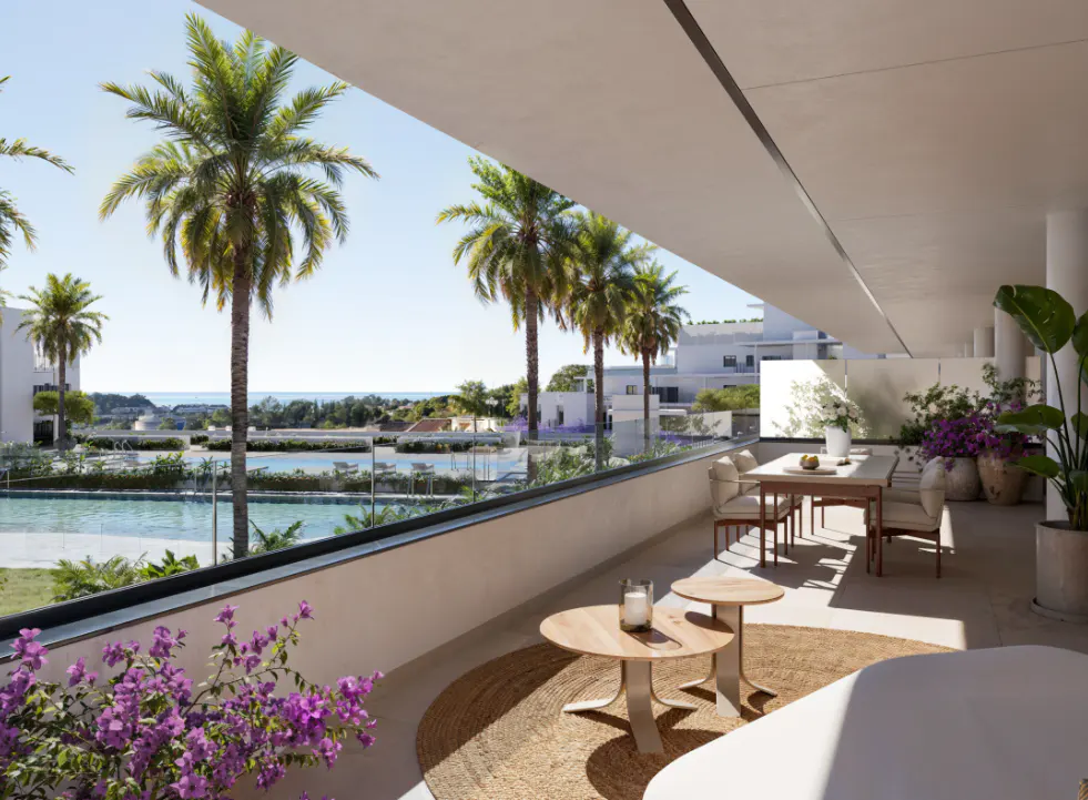 Balcony view with palm trees, ocean, and outdoor furniture. Table with chairs, round tables, and plants create a relaxing space.