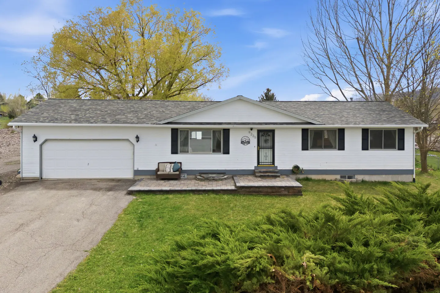 A white single-story house with a gray roof, black shutters, and a one-car garage. A green lawn and trees are in the foreground and background.