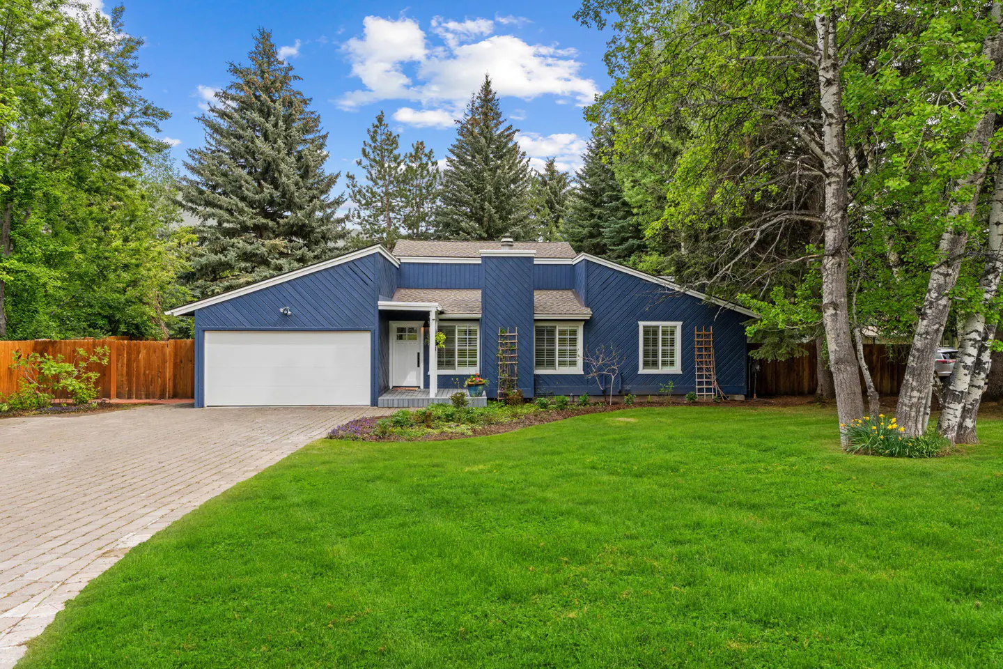 Blue single-story house with white trim, garage, and green lawn. Trees surround the property under a partly cloudy sky.