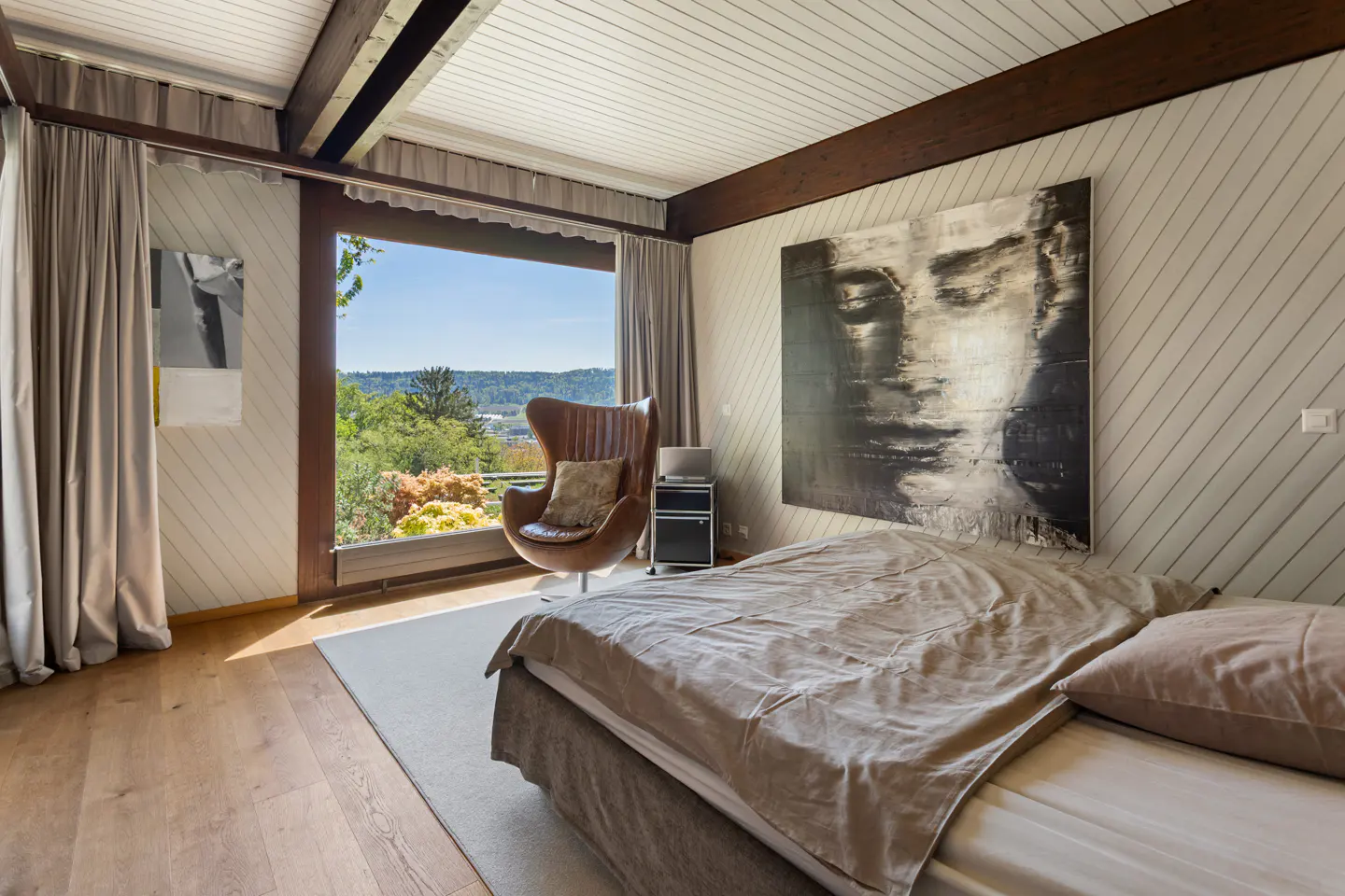 Bedroom with wood floors, a large window with a view, and a brown leather chair. A large black and white painting hangs above the bed.