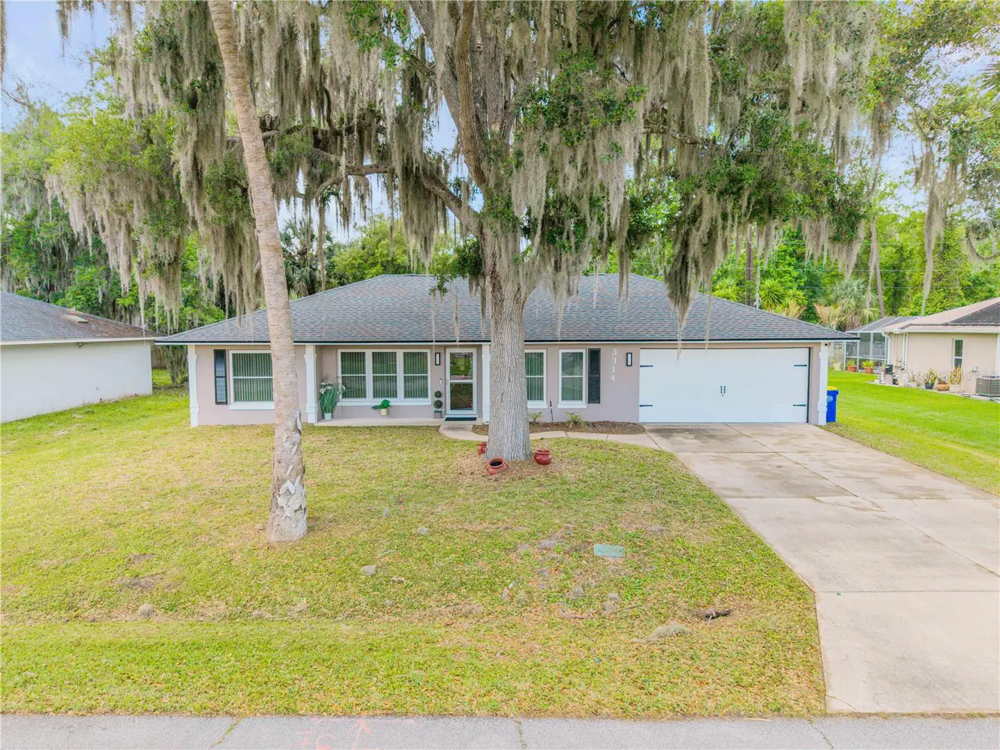 Single-story home with a gray roof, light pink exterior, and white garage door. A large tree with Spanish moss dominates the front yard.