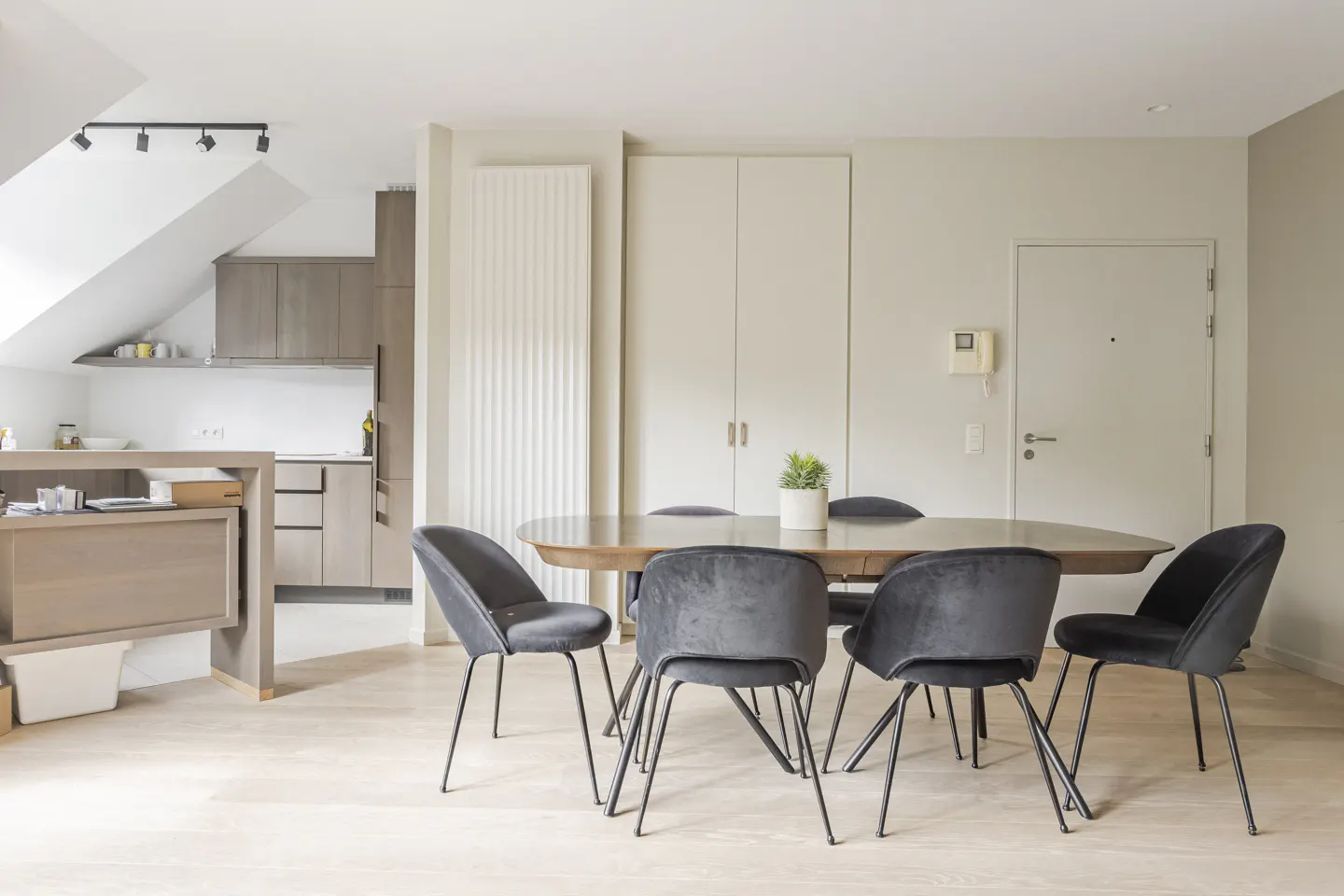 A modern dining area with a wooden table, black velvet chairs, and a small potted plant. The kitchen is visible in the background.