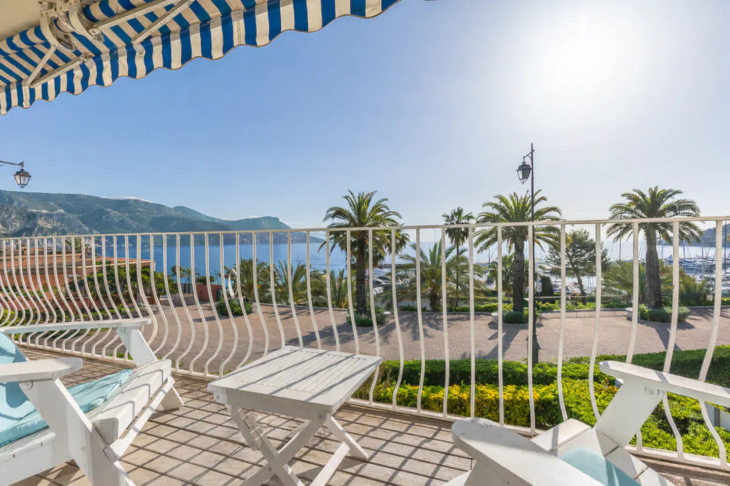 Balcony view with white chairs, table, and railing overlooking palm trees, a marina, and the ocean under a blue and white striped awning.
