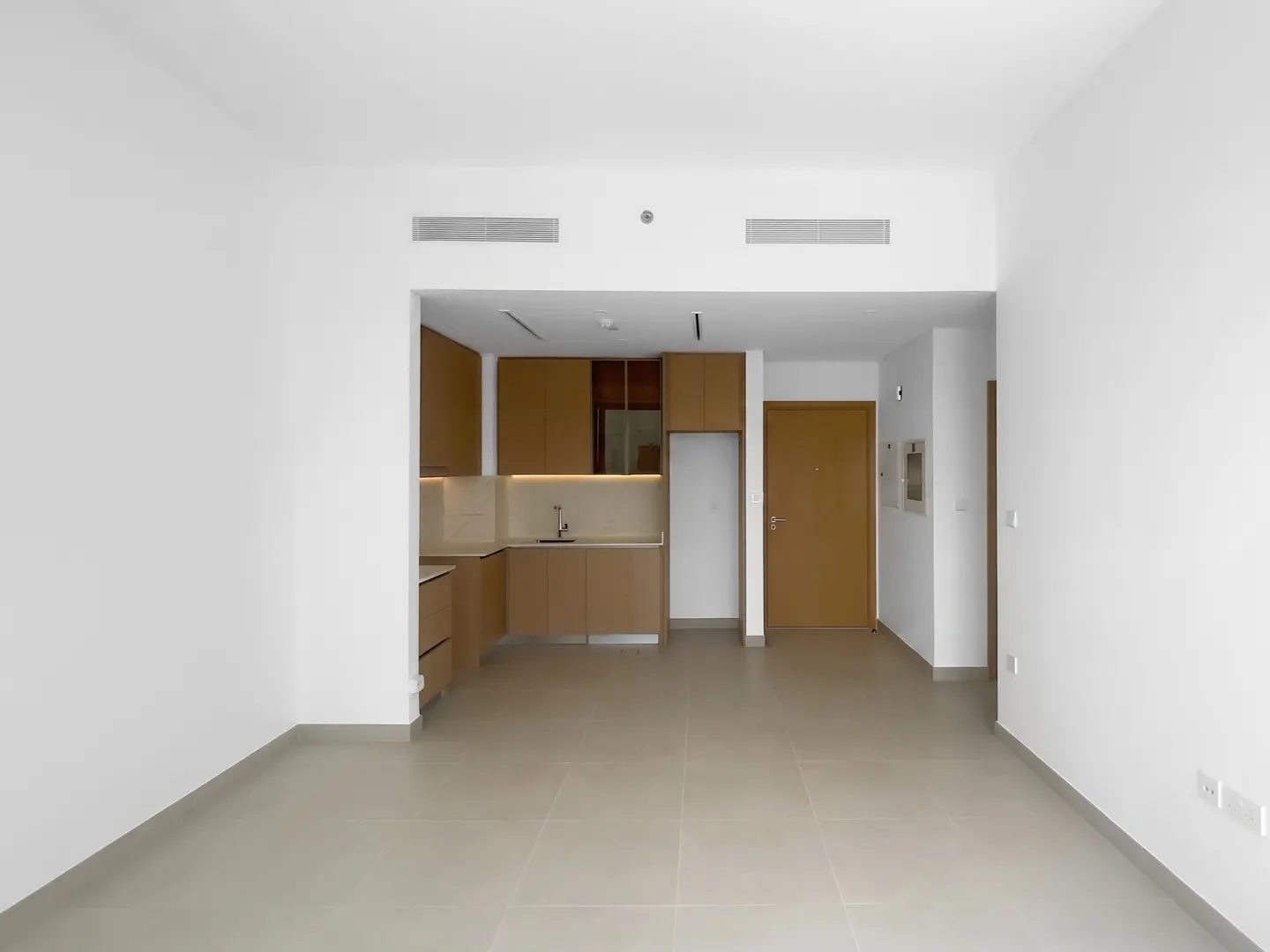 An empty apartment with white walls, beige tile floors, and a brown kitchen with a sink and cabinets. A brown door is on the right.