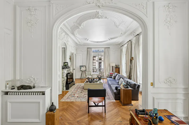 Elegant living room with ornate white moldings, herringbone wood floors, a grey sofa, and a large arched doorway.