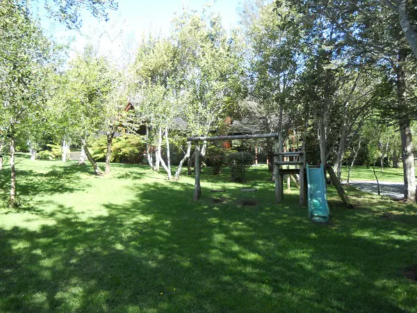 Lush green lawn with trees, a wooden swing set, and a blue slide. A house is visible in the background.