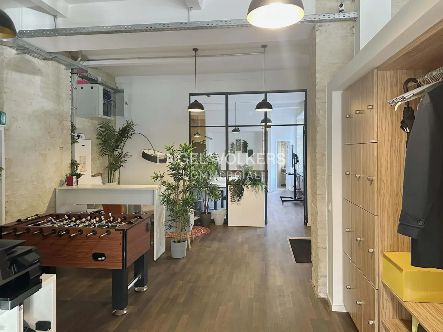 Office space with a foosball table, plants, and wooden lockers. Black framed glass doors lead to another room. Overhead lighting is visible.