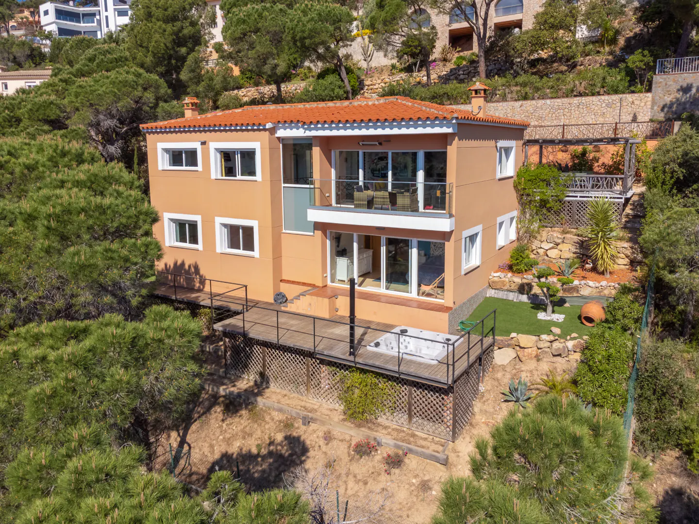Two-story peach house with a red tile roof, surrounded by trees. A wooden deck with a hot tub is in the foreground.