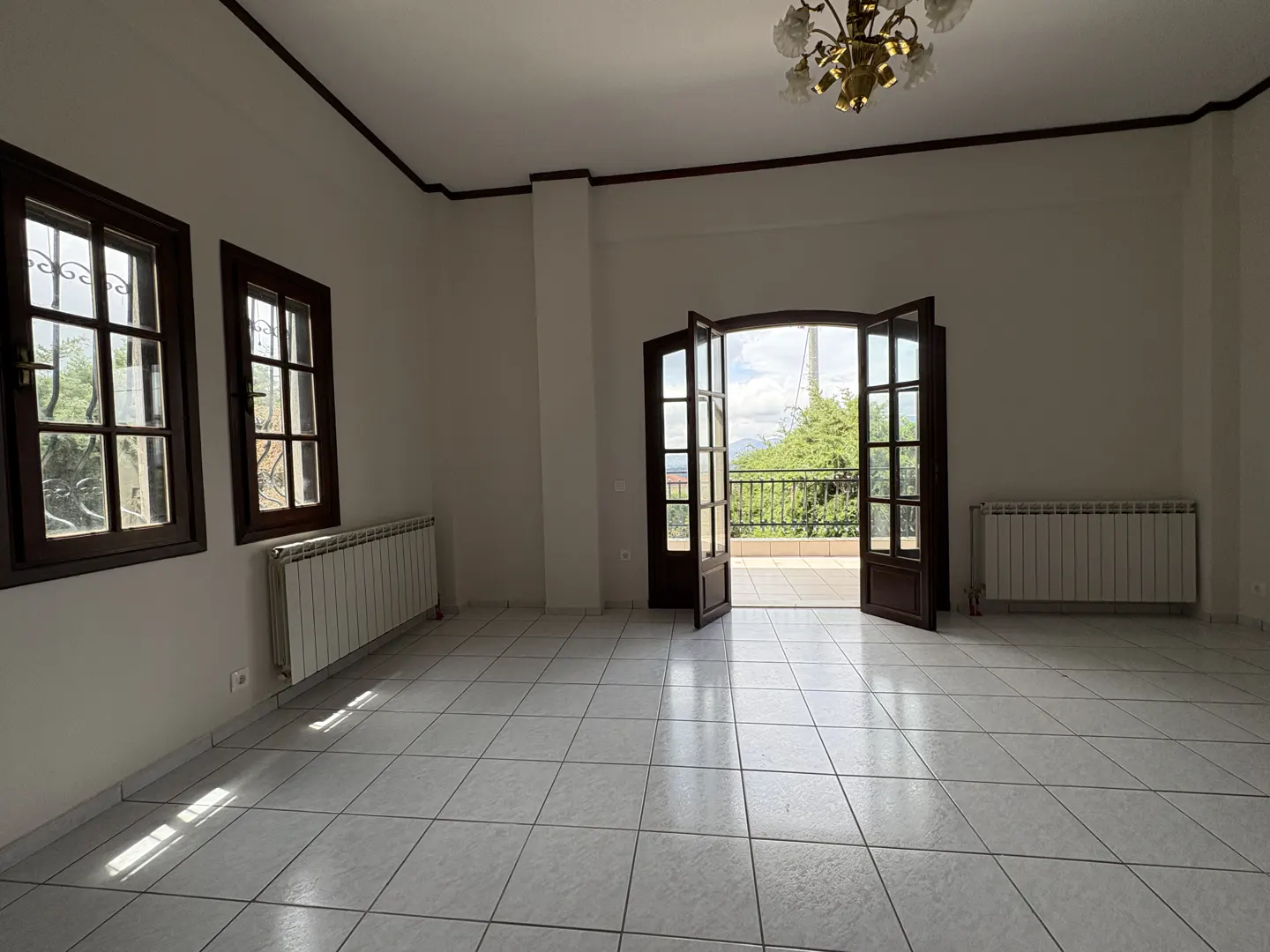 Bright, empty room with white tile floor, two windows, and open doors to a balcony with trees and sky visible.