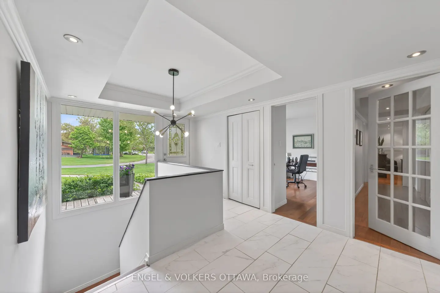 Bright, white foyer with marble floors, a modern chandelier, and a large window overlooking a green lawn. Doors lead to an office and another room.