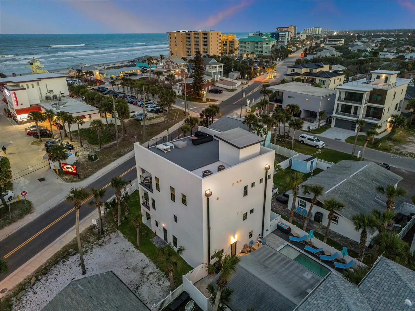Aerial view of a modern, white, multi-story home with a rooftop deck near a beach and coastal town.