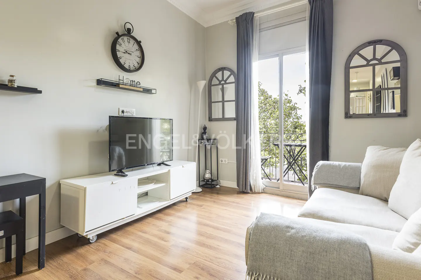 Living room with a white sofa, TV on a white stand, and balcony with trees visible. Gray curtains frame the balcony door.