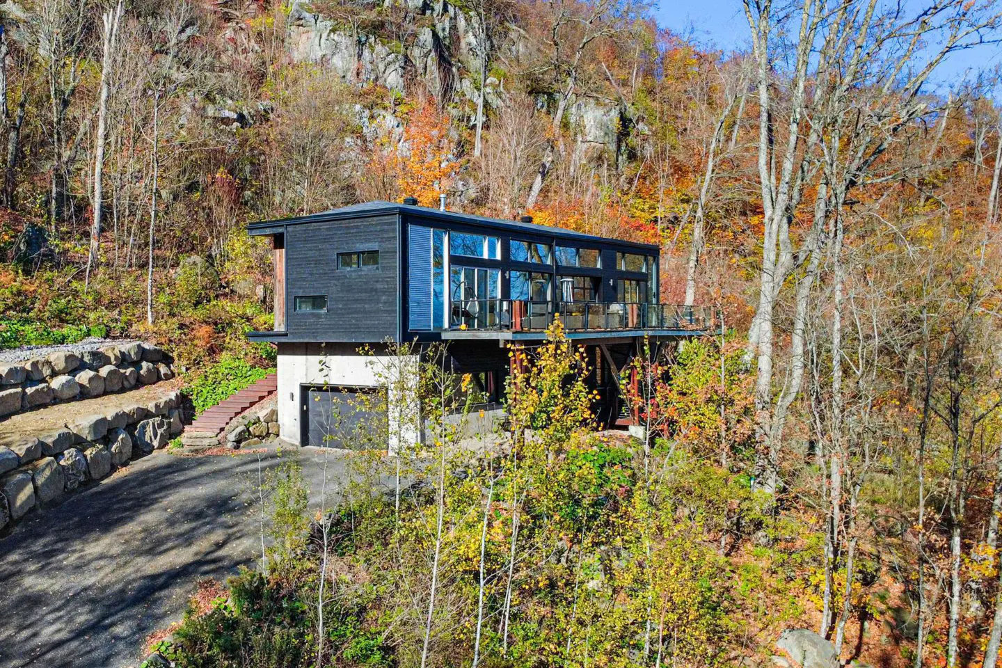 Modern house with black siding and large windows, built on a hillside with fall foliage. A driveway leads to a garage.