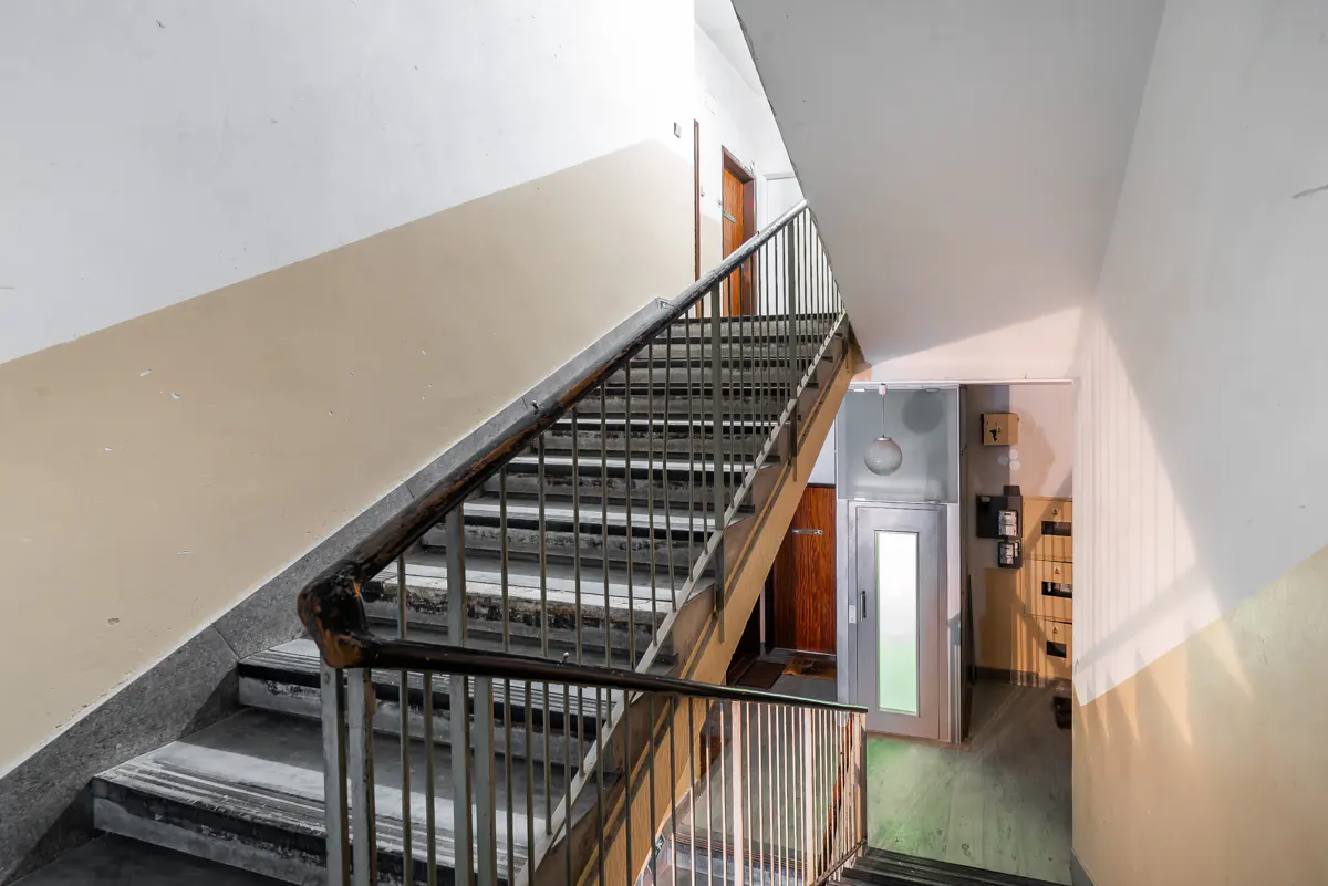 Interior view of a stairwell with gray stairs, metal railings, and a vintage elevator. Walls are painted white and beige.