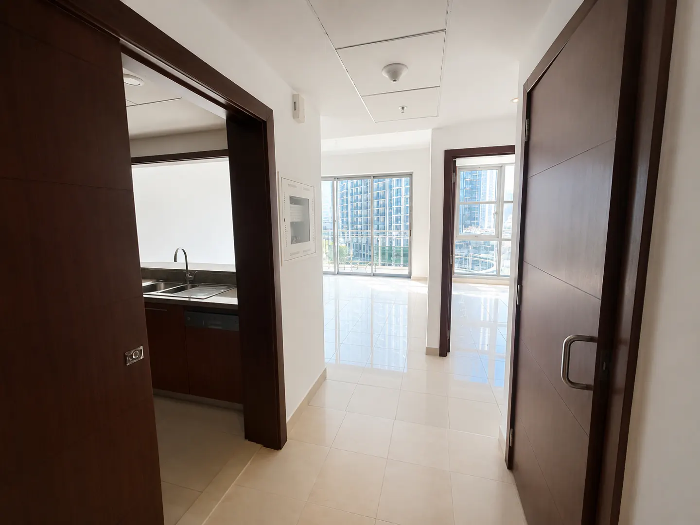 Interior view of a modern apartment with beige tile floors, brown doors, and a glimpse of a kitchen and city view.