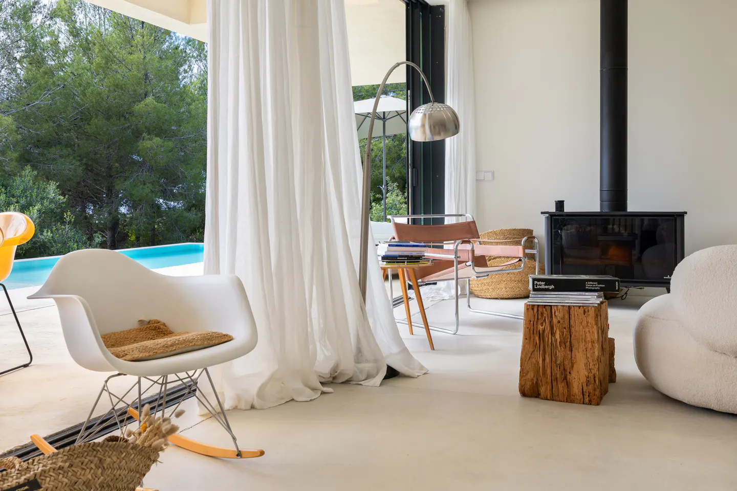 Bright living room with a white rocking chair, a leather chair, a black fireplace, and a view of a pool and trees.