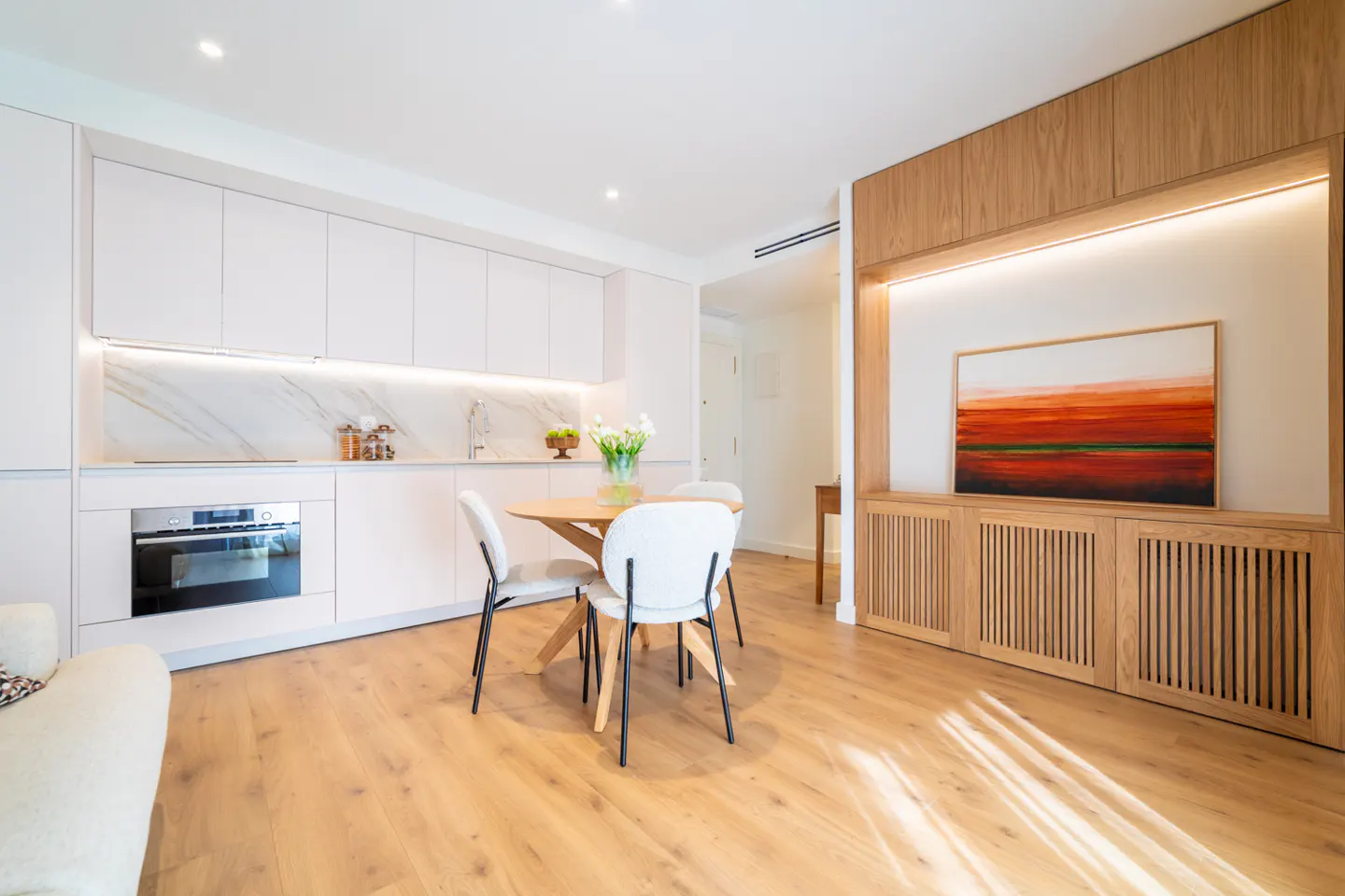 Bright, modern apartment interior with white cabinets, wood floors, and a round table with white chairs. An abstract painting hangs on the wall.