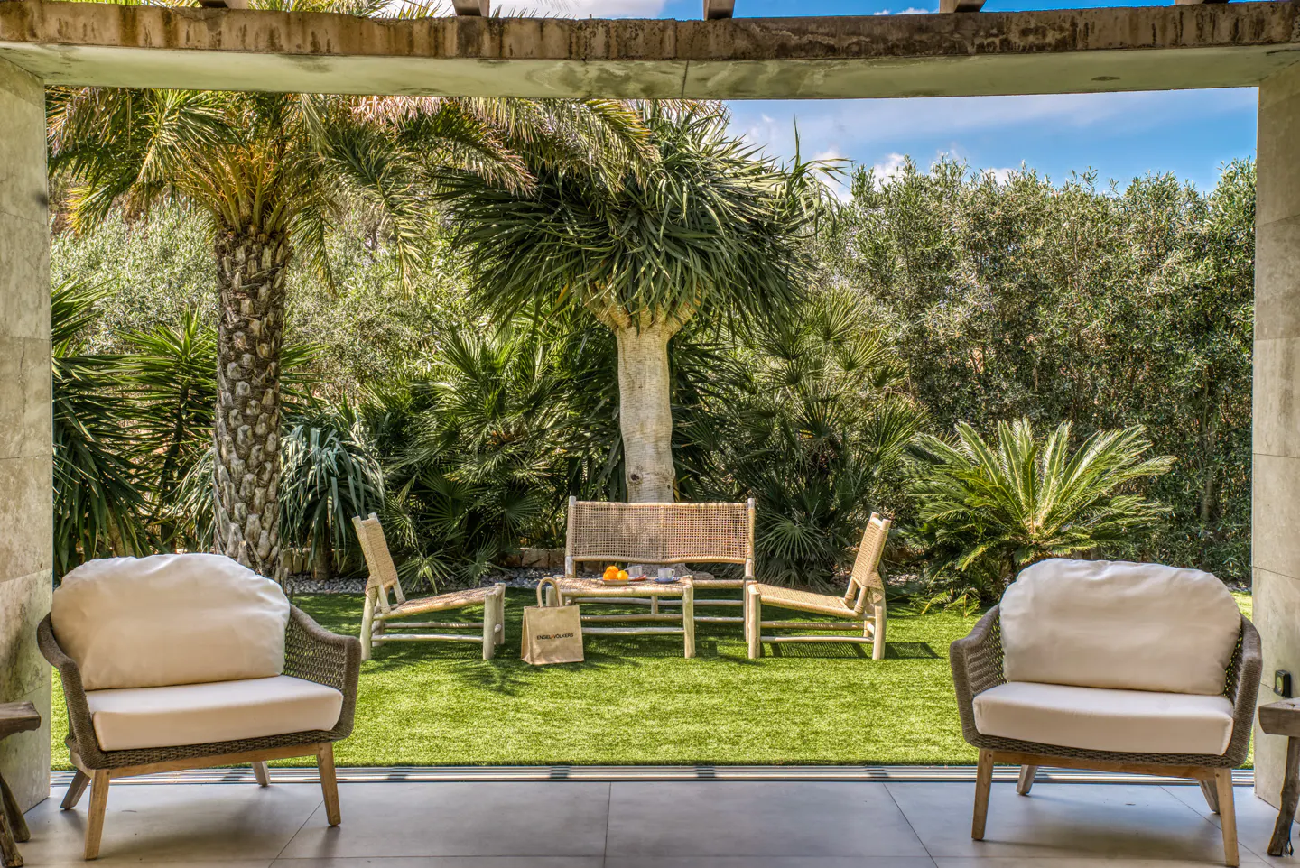 Outdoor patio with two armchairs and a table with two chairs on a green lawn, surrounded by lush trees and plants.