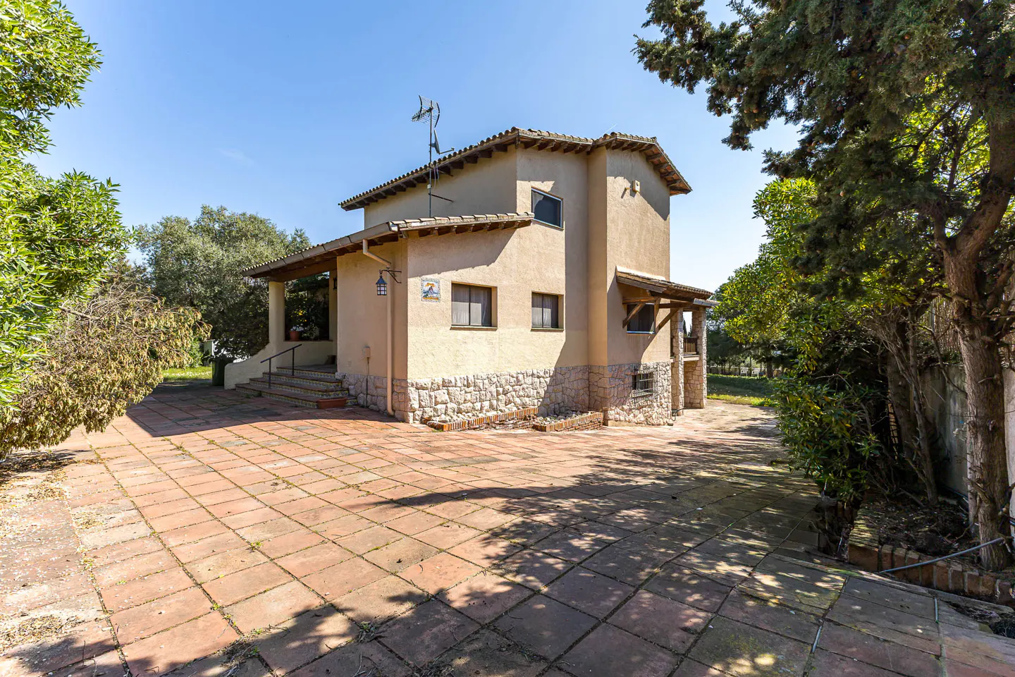 Two-story beige house with stone base, brown tile roof, and brick patio. Trees surround the house under a blue sky.