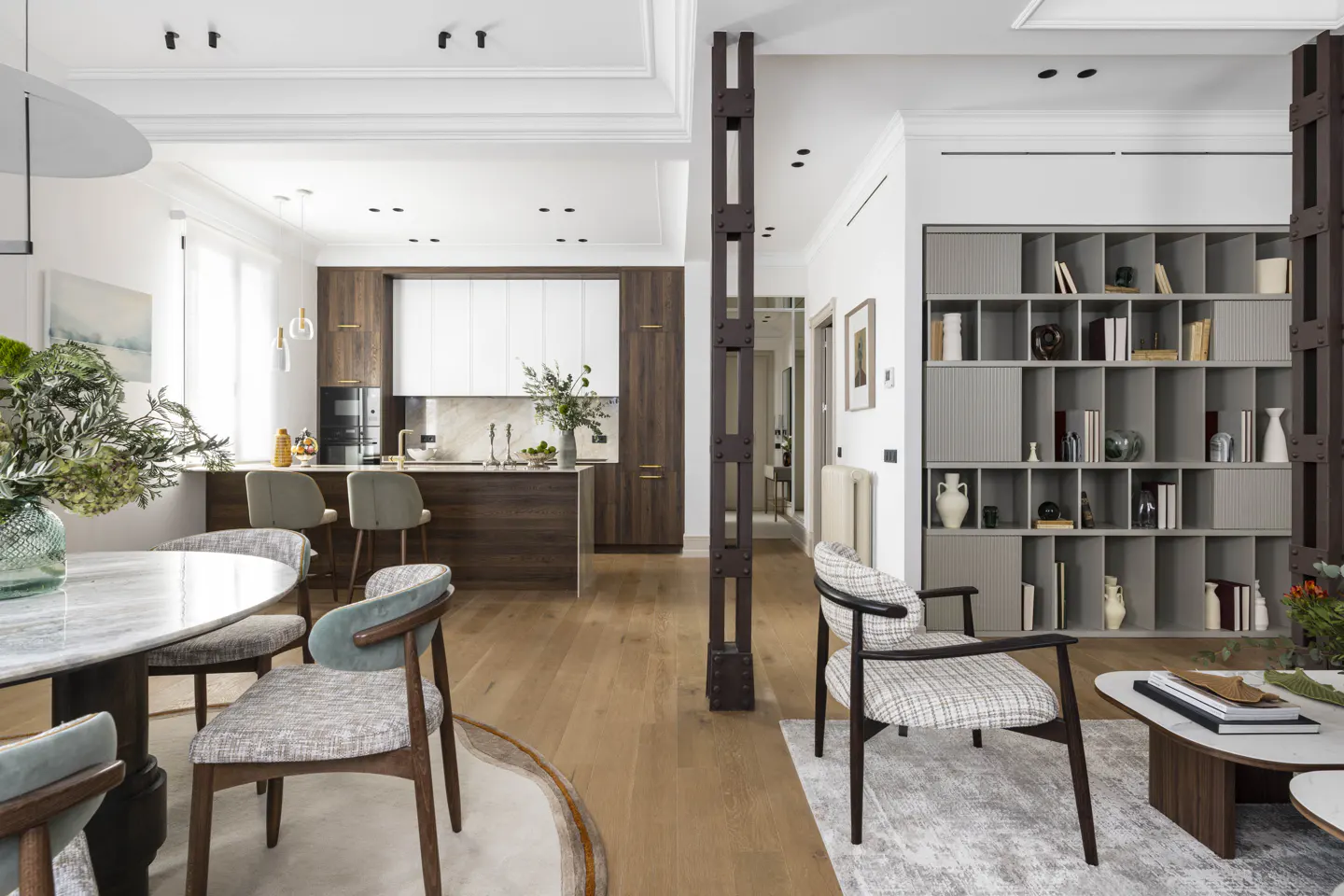 Open-concept living space with wood floors, a kitchen island, dining table, and gray bookcase. Natural light fills the room.