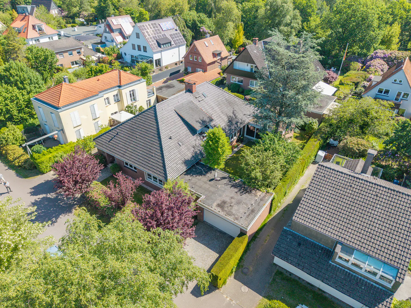 Aerial view of a suburban neighborhood featuring a central house with a dark tiled roof, garage, and lush green trees.